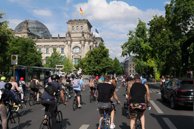 Eine Gruppe von Menschen, die auf Fahrrädern eine von Bäumen gesäumte Straße in Berlin, Deutschland, entlangfahren, mit Gebäuden, einer Bushaltestelle auf der rechten Seite und einer Flagge, die auf einem der Gebäude weht, unter einem bewölkten Himmel.